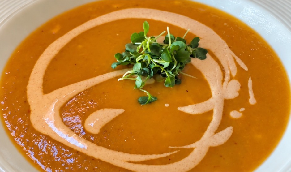 Orange soup served in a white plate with a cream swirl and fresh microgreens